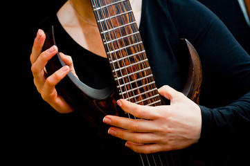 Seven-string electric guitar made of dark wood and human hands, shot on a dark background....