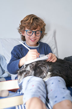 blond boy with glasses writing in a notebook sitting on the bed with his dog