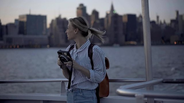 Happy cheerful female tourist in casual wear photographing scenic views during voyage trip near Manhattan island in New York, smiling amateur with instant camera enjoying water boat excursion
