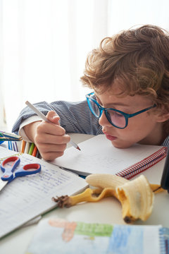 Boy with curly hair sitting on sofa near round table and writing in notebook while doing homework in cozy living room at home