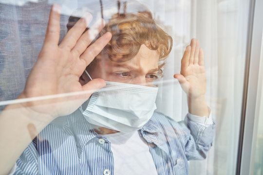 Boy in medical mask keeping hand on glass and looking through window while staying home during quarantine
