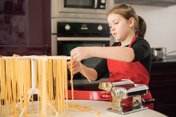 Serious preteen girl in red apron hanging raw noodle on rack while standing at table with pasta machine in kitchen