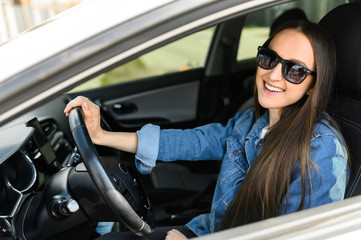 Fototapeta na wymiar A young girl driving a car. She is in casual jeans jacket and in sunglasses looks at camera and smiles