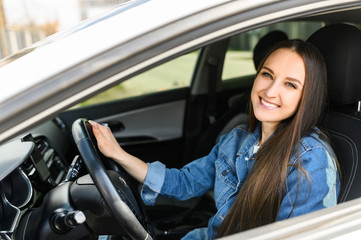 A young girl driving a car. She is in casual jeans jacket looks at camera and smiles