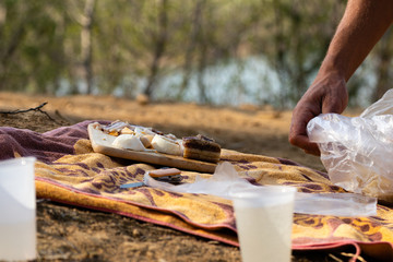 Man setting up picnic with home made pancetta, home bottled wine, eggs and home made breed on the towel in the nature near the lake, with plastic glasses in the foreground.  