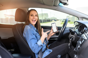 A young beautiful woman driving a car, she points to blank phone screen and smiles. Mobile apps for driver. Inside car view