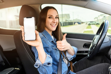 A young happy woman driving a car, she shows empty phone screen and thumb up. Useful mobile apps...