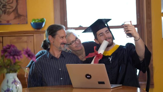 Mother and father smile as online graduate son raises phone and takes a selfie of them in front of computer with diploma during virtual commencement ceremony.