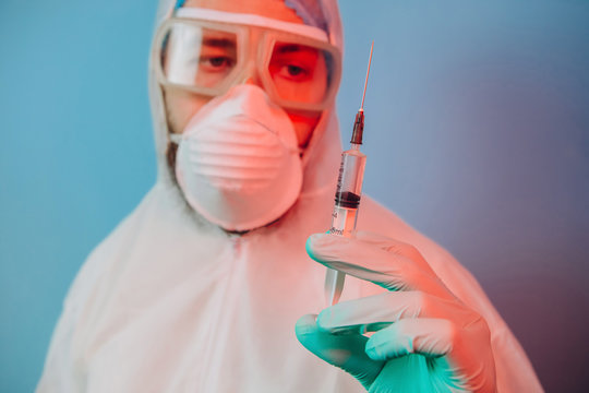 Doctor In Protective Uniform, Reperitor, Glasses, Gloves On Blue Background In Neon Light. Close-up Portrait Of Doctor In Red Neon. Tired Man Is Battling Coronavirus. Syringe With Vaccine. Covid 19