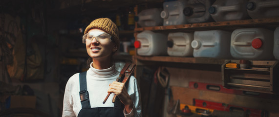 Portrait of smiling young builder girl standing in old workshop room, wearing construction glasses.