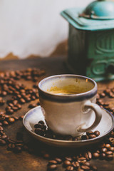 Granos de cafe por la mesa de madera rústica. Un expresso en una taza blanca. De fundo se ve un moledor. La imagen es acogedora, caliente, vintage.