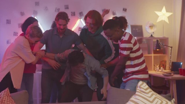 Bored man sitting on couch with bottle of beer while joyous friends pulling him up and dancing together at home party in room decorated with neon light