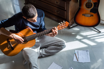 Obraz premium Man playing guitar and composing music at home near a bright window on a sunny day. Casual musician sitting on the floor playing the guitar.