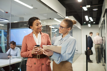 Two young businesswomen discussing documents and drinking coffee during working day at office