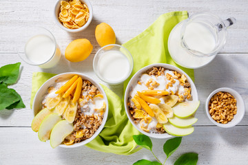 Granola and vegetarian yogurt with slices of apple, apricot, banana and a jug of milk on a white wooden background. Healthy breakfast concept. Top view