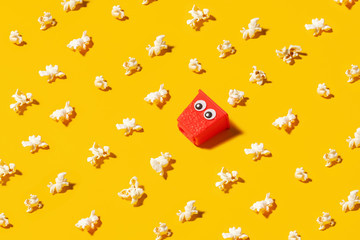 Mini red popcorn box carton with googly eyes on a yellow background surrounded by popped popcorn.