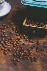 Granos de cafe por la mesa de madera rústica. Un expresso en una taza blanca. De fundo se ve un moledor. La imagen es acogedora, caliente, vintage.