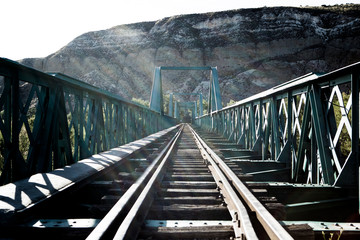 Vía de tren sobre puente con destellos de luz y montaña al fondo