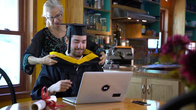 As part of a virtual commencement ceremony, a mother hooding her son graduating in front of laptop computer in their home dining room.