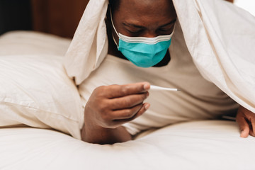 African American male in blue and white surgical mask laying in bed looking at a thermometer, displaying signs of being sick