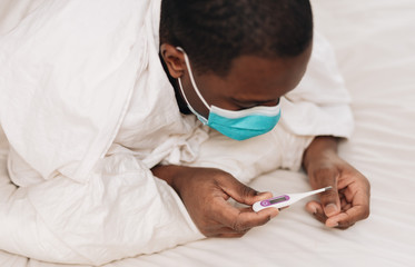 African American male in blue and white surgical mask laying in bed looking at a thermometer, displaying signs of being sick
