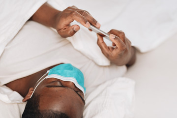 Obraz na płótnie Canvas African American man laying in bed with blue and white surgical face mask looking at cellphone, displaying signs of sickness