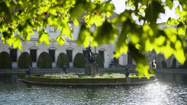 Wallenstein Garden in sunny spring day. View through fresh leaves on foreground bright sun reflectingglare in water, lake with statues and fountain, famous sight of Prague 