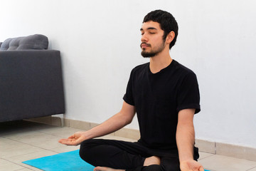 young hispanic man wearing black sportswear meditating in a yoga session in the lotus flower pose , at home