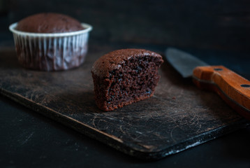 Delicious cutaway chocolate muffin on a dark old wooden board next to a knife and a paper muffin.