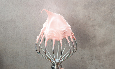 Pink whipped swiss meringue cream on top of large metal beater. Plain grey background.