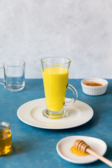 Tall glass with herbal,traditional Indian drink with turmeric stands on white flat plate. Golden milk for disease prevention and immune support.Next to it are ingredients curcum and honey. Copy space