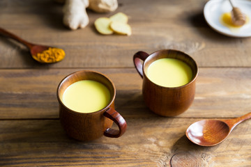Obraz na płótnie Canvas Herbal,healthy,Indian drink golden milk with turmeric root powder immune booster in two wooden cups stand on a background. Next to it are two wooden spoons and the ingredients honey,ginger and curcuma
