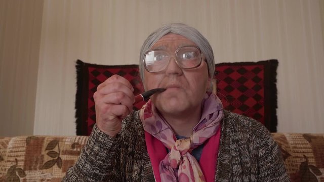 Funny GRANNY with eccentric look smokes a TOBACCO PIPE and reacts with surprise &mdash; comic close-up in retro home interior.