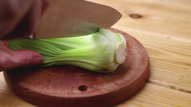 Male hand cuts fresh green bok choy or pac choi chinese cabbage with a knife on a brown wooden kitchen board. Side view, close up.