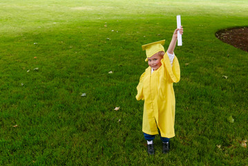 Young boy in a yellow cap and gown smiles and holds up his preschool graduation diploma on a green grass field