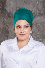 Portrait of attractive young female doctor in white medical jacket and  green medical cap on a grey background.