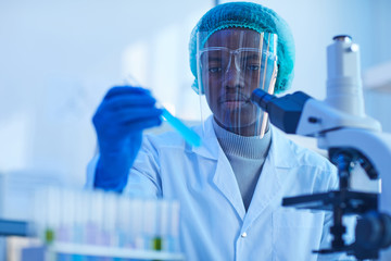 African scientist in protective gloves holding test tube with blue liquid while working with...