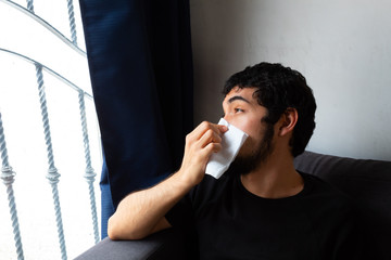 Young Hispanic man with a beard showing discomfort, pain, illness, suffering from coronavirus symptoms and confinement, covering his mouth and nose with a tissue