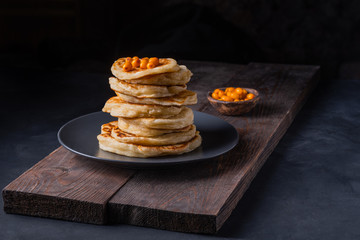 A stack of sweet pancakes with sea buckthorn berries and honey on a dark background in a low key