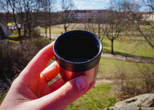 Female hand holding a pink thermos mug in a sunny day in a park drinking hot tea or hot coffee outdoors