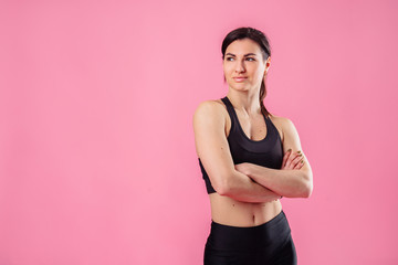 Young pumped up girl coach posing in the studio on a pink background. Dressed in a black top and black pants. Feeling very relaxed and natural, smiles broadly and holds hands on the waist