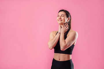 Obraz premium Slim pumped up girl fitness trainer stands against pink background in the studio in black sportswear. She hugs herself with hands, closing her eyes, smiling, dreaming, and raising her head up