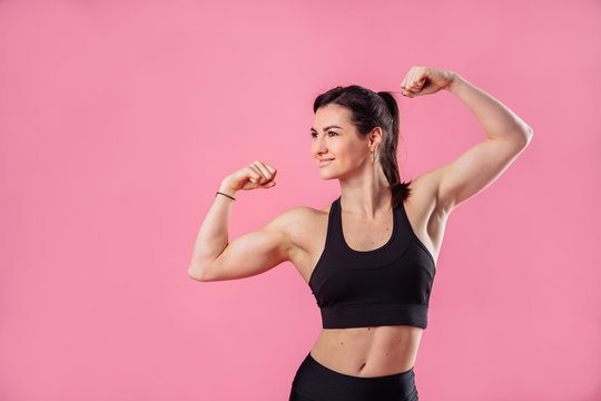 Half lenght portrait of beautiful, pumped up girl in a black tight-fitting top and leggings smiling and showing her big, pumped up muscles in her arms, isolated on pink background in photostudio © Daria