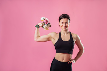 Funny photo of a young muscular fitness girl with a bouquet in her hands, that sniffs flowers and pumps up muscles. Cheerfully smiling european sporty woman, demonstrating biceps on pink background