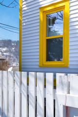 A white and yellow building with a single double hung window with yellow trim. There's a white wooden picket fence in the foreground made of textured wood. The background has houses, mountain and tree
