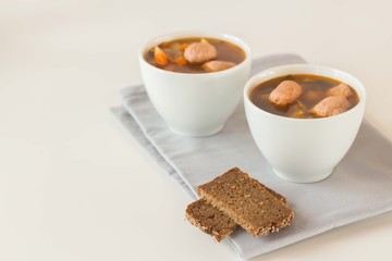 A bowl of soup with sausages on a gray napkin, next to two slices of brown bread
