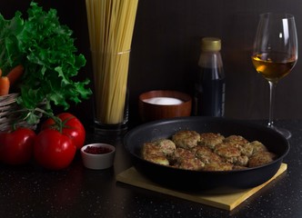 Meatballs in a pan are on the table, next to tomatoes are green salad and carrots in a basket, a glass of white wine, sauce in a bottle, spaghetti in a jar, spices and salt
