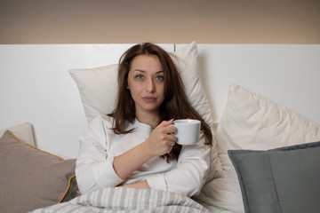 Calm beautiful sexy woman in casual wear sitting in bed with cup of tea in hand, calmly and confidently looking at camera. Weekend, relax, bedtime, home self-isolation and quarantine concept