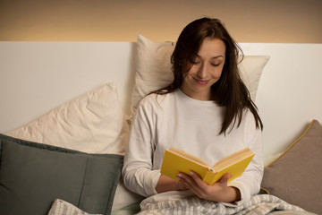 pretty cute dark-haired girl in pajamas smiling while reading book with yellow cover on lazy day modern interior backlighting of bed headboard