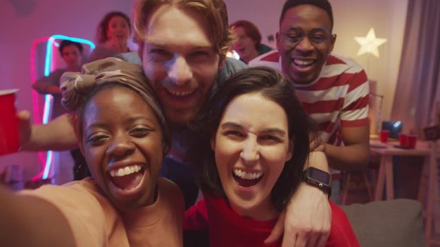 Young multi-ethnic friends happily smiling, embracing and looking at camera while taking group selfie at home party in living room with neon decorations